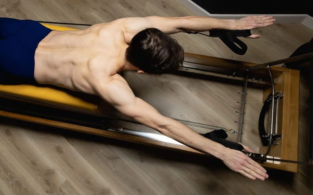 Muscular man lying face down on a wooden machine demonstrating the upper body benefits of Pilates Reformer workouts.